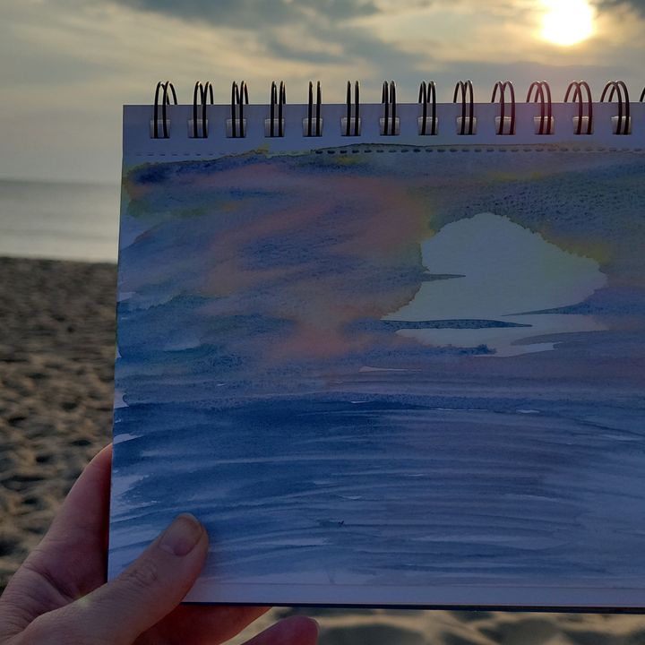 Hand holding a watercolor sketchbook painting of a sunrise on a beach.