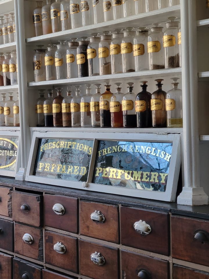 A wall with drawers, apothecary bottles, and signs saying Prescriptions Prepared and French and English Perfumery